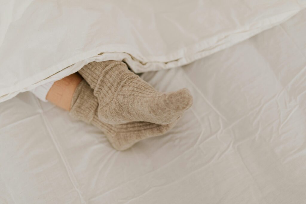 Comfortable feet in woolen socks peek out under a soft white linen blanket on a bed.